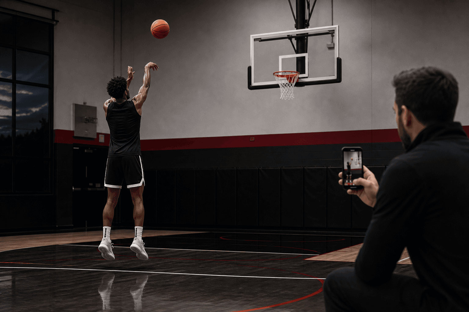 Basketball player shooting in a local indoor gym while a trainer records the rep on a phone.