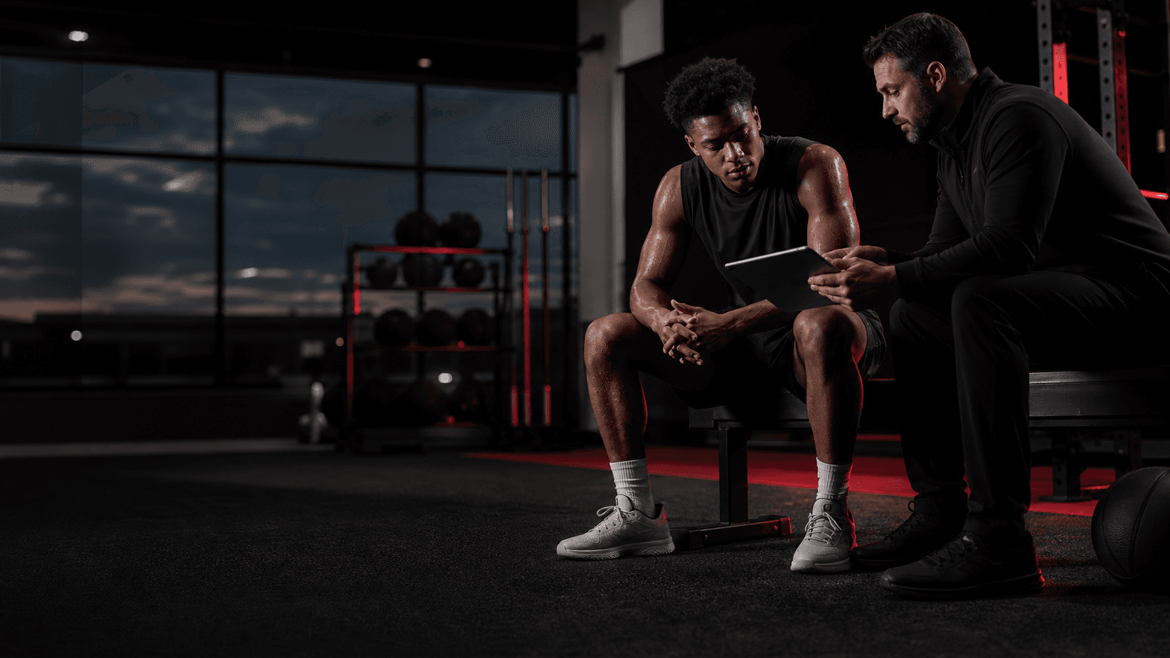 Athlete seated between reps while a coach reviews practice data on a tablet in a dark training facility.